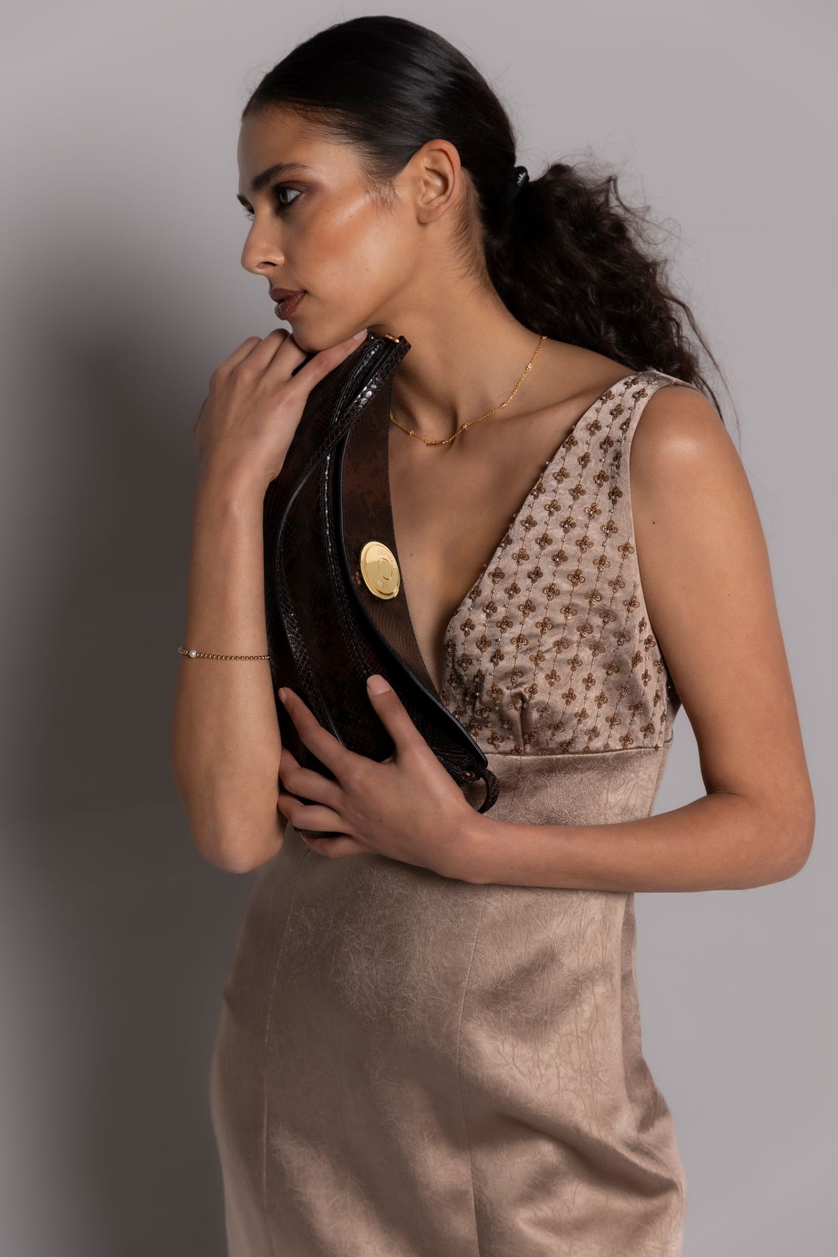 Model in beigem Kleid mit dunkler Clutch vor grauer Wand